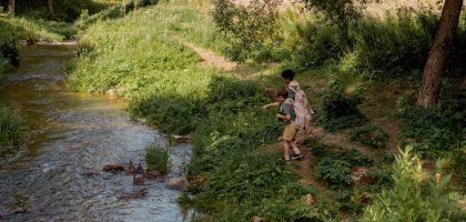 Two boys in scouting uniforms explore a forest path by a stream, enjoying nature.